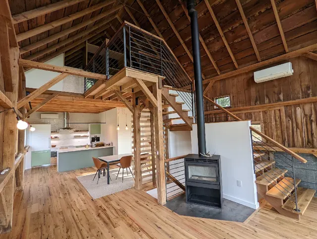A timber interior with exposed beams, wood flooring, a loft railing, stairs, and a wood stove.